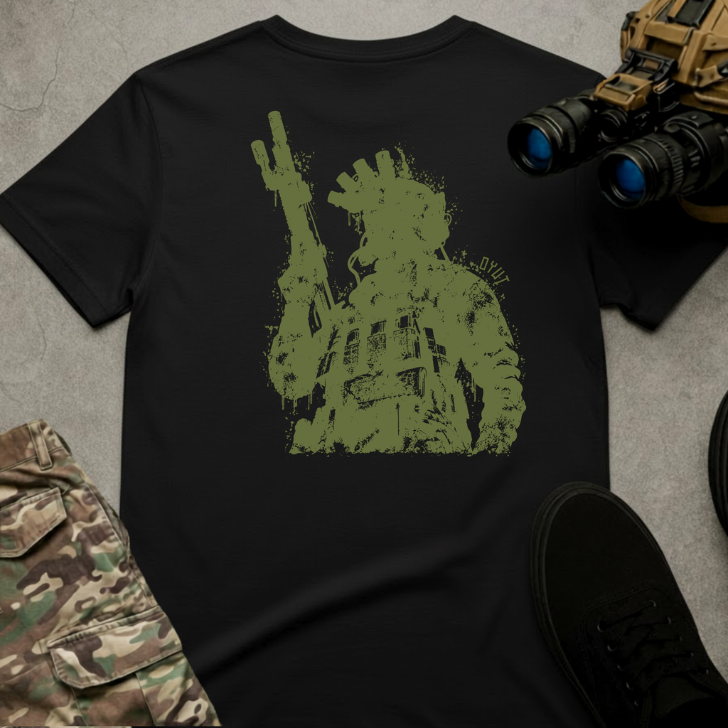 A black short-sleeve graphic T-shirt laid flat on a gray textured surface, surrounded by tactical-style props including camouflage pants, black shoes, and binoculars. The shirt features a large distressed olive-green graphic of a tactical soldier wearing combat gear and holding a rifle, creating a rugged military-inspired design.