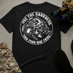 A black short-sleeve graphic T-shirt laid flat on a gray textured surface, surrounded by tactical-style props including camouflage pants, black shoes, and night-vision binoculars. The shirt features a large distressed white circular graphic of a skull wearing a tactical helmet and multi-lens night-vision goggles, with the text “USE THE DARKNESS” above and “OWN YOUR WAY TRIBE” below.