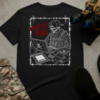 A black short-sleeve graphic T-shirt laid flat on a gray textured surface, surrounded by tactical-style props including camouflage pants, black shoes, and night-vision binoculars. The shirt features a distressed white and red graphic of a skull-faced tactical figure wearing a backward cap and tactical vest while working on a laptop, surrounded by electronic equipment and gear. Bold red text reads “HUNT PEDOS” beside the figure.
