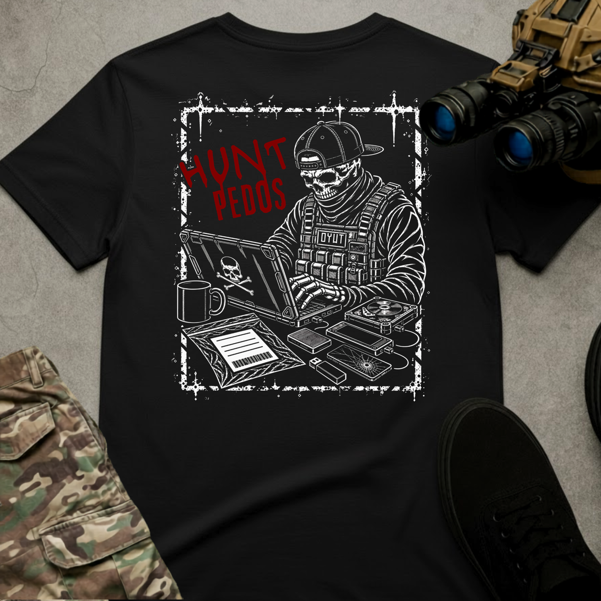 A black short-sleeve graphic T-shirt laid flat on a gray textured surface, surrounded by tactical-style props including camouflage pants, black shoes, and night-vision binoculars. The shirt features a distressed white and red graphic of a skull-faced tactical figure wearing a backward cap and tactical vest while working on a laptop, surrounded by electronic equipment and gear. Bold red text reads “HUNT PEDOS” beside the figure.