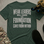 A dark heather green t-shirt with a distressed white graphic text reading 'WEAK LEADERS WILL CAUSE THE FOUNDATION TO CAVE FROM WITHIN', presented in a flat-lay arrangement with tactical boots, binoculars, and camouflage pants on a gray concrete background.