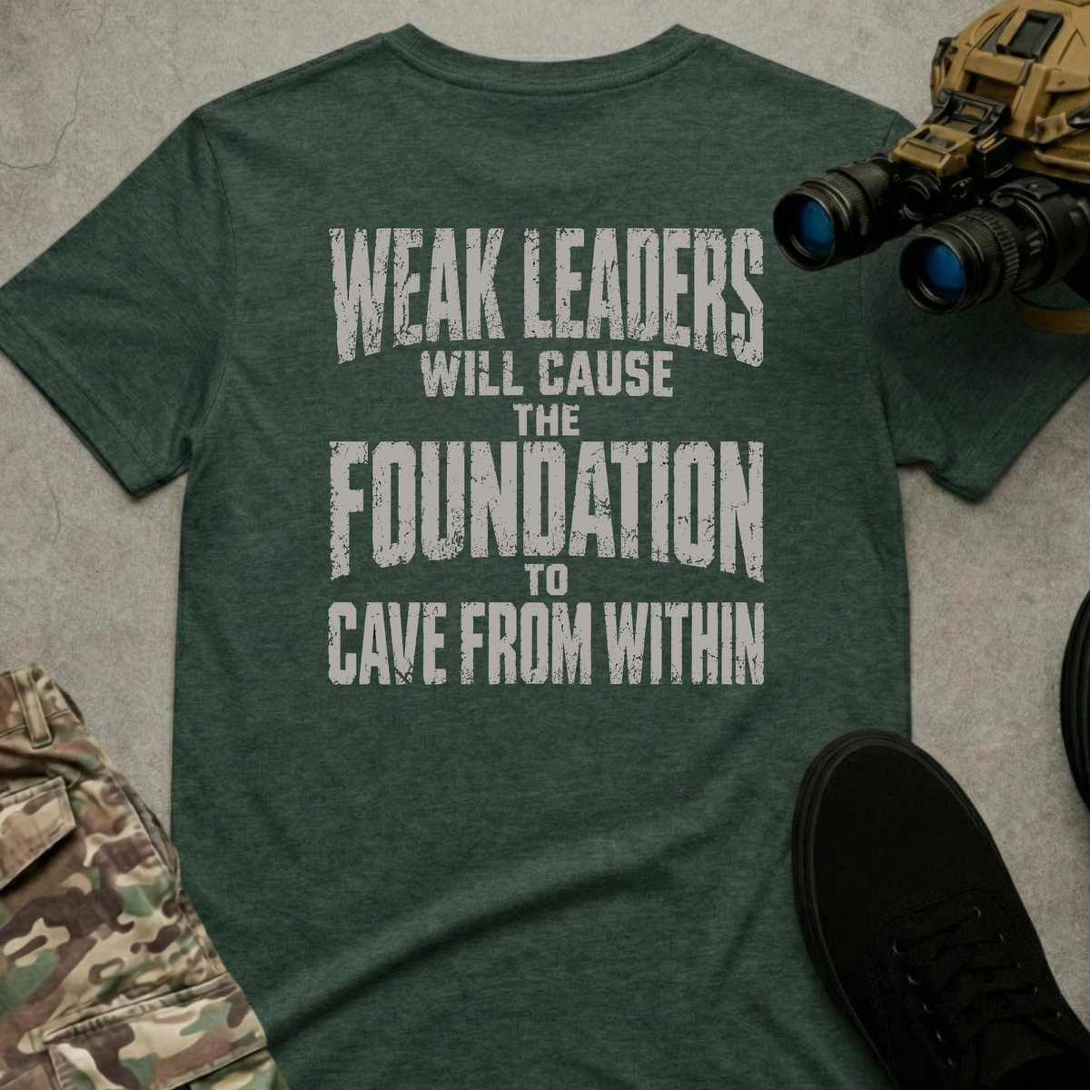 A dark heather green t-shirt with a distressed white graphic text reading 'WEAK LEADERS WILL CAUSE THE FOUNDATION TO CAVE FROM WITHIN', presented in a flat-lay arrangement with tactical boots, binoculars, and camouflage pants on a gray concrete background.