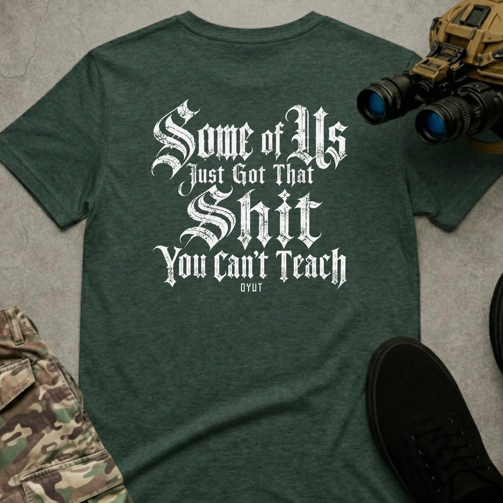 A dark heather green t-shirt with a white gothic-style text graphic reading 'Some of Us Just Got That Shit You Can't Teach (GYUT)', presented in a flat-lay arrangement with tactical gear props on a gray concrete background.