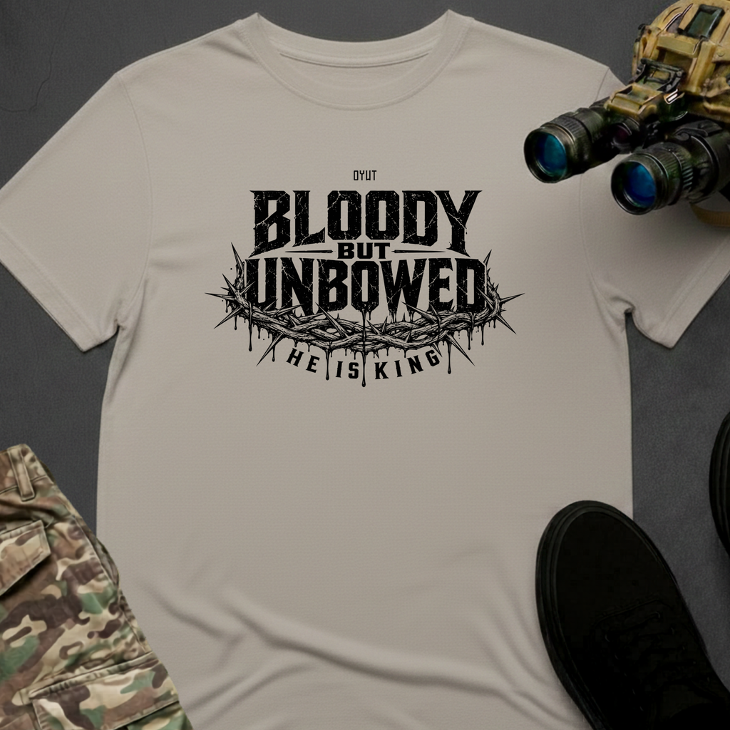 A light beige crewneck t-shirt featuring a black graphic print that reads 'BLOODY BUT UNBOWED, HE IS KING.' The shirt is styled in a flat-lay arrangement with tactical gear props, including binoculars, a black shoe, and camouflage pants, on a dark gray background.