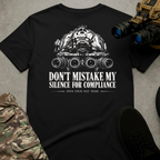 A black short-sleeve graphic T-shirt laid flat on a gray textured surface, featuring a white military-style tank illustration and the text 'DON'T MISTAKE MY SILENCE FOR COMPLIANCE' with a smaller line 'OWN YOUR WAY THERE.' Tactical boots, binoculars, and camouflage pants are placed around it as styling props.