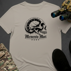 A light beige crewneck t-shirt laid flat on a dark gray background, featuring a black graphic of a skull with a clock face inside its eye socket and the gothic text 'Memento Mori' and 'OWE'. Tactical gear props, including binoculars, camouflage pants, and a black shoe, are arranged around the shirt.