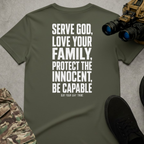 An olive green short-sleeve t-shirt with bold white text reading 'SERVE GOD, LOVE YOUR FAMILY, PROTECT THE INNOCENT, BE CAPABLE' and smaller text 'OUR DAY ONE' below, shown in a styled flat-lay with camouflage pants, black shoes, and binoculars on a gray textured background.