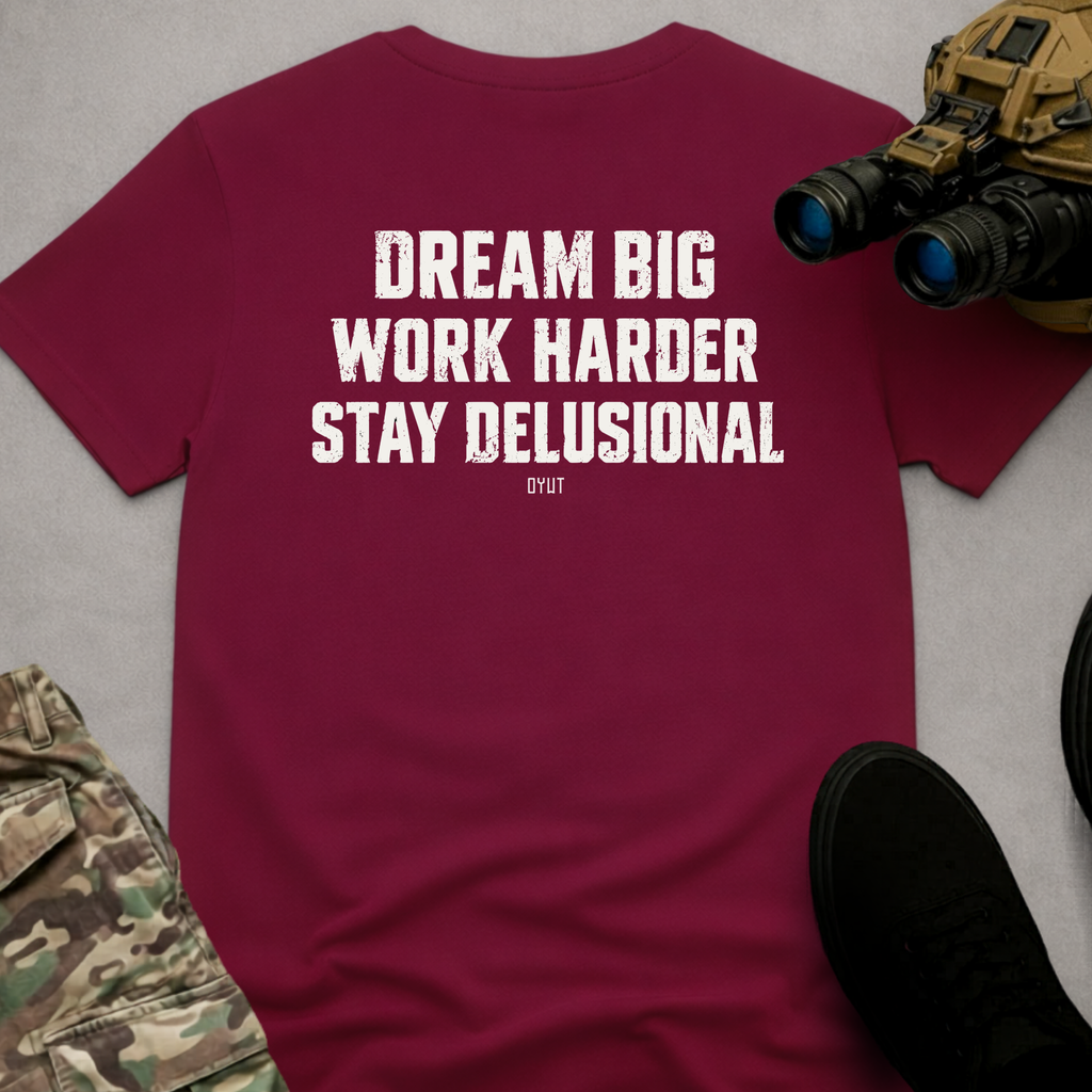 A burgundy short-sleeve t-shirt with a white distressed text graphic reading 'DREAM BIG WORK HARDER STAY DELUSIONAL' and 'DYOT' underneath, styled in a flat-lay with tactical gear props on a gray background.
