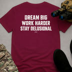 A burgundy short-sleeve t-shirt with a white distressed text graphic reading 'DREAM BIG WORK HARDER STAY DELUSIONAL' and 'DYOT' underneath, styled in a flat-lay with tactical gear props on a gray background.