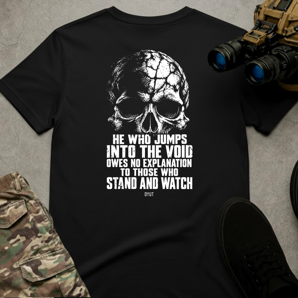 A black short-sleeve graphic T-shirt laid flat on a gray textured surface, featuring a large white skull and distressed text that reads, 'HE WHO JUMPS INTO THE VOID OWES NO EXPLANATION TO THOSE WHO STAND AND WATCH' with 'OG197' below. Tactical-style props including binoculars, camouflage pants, and black shoes surround the shirt.