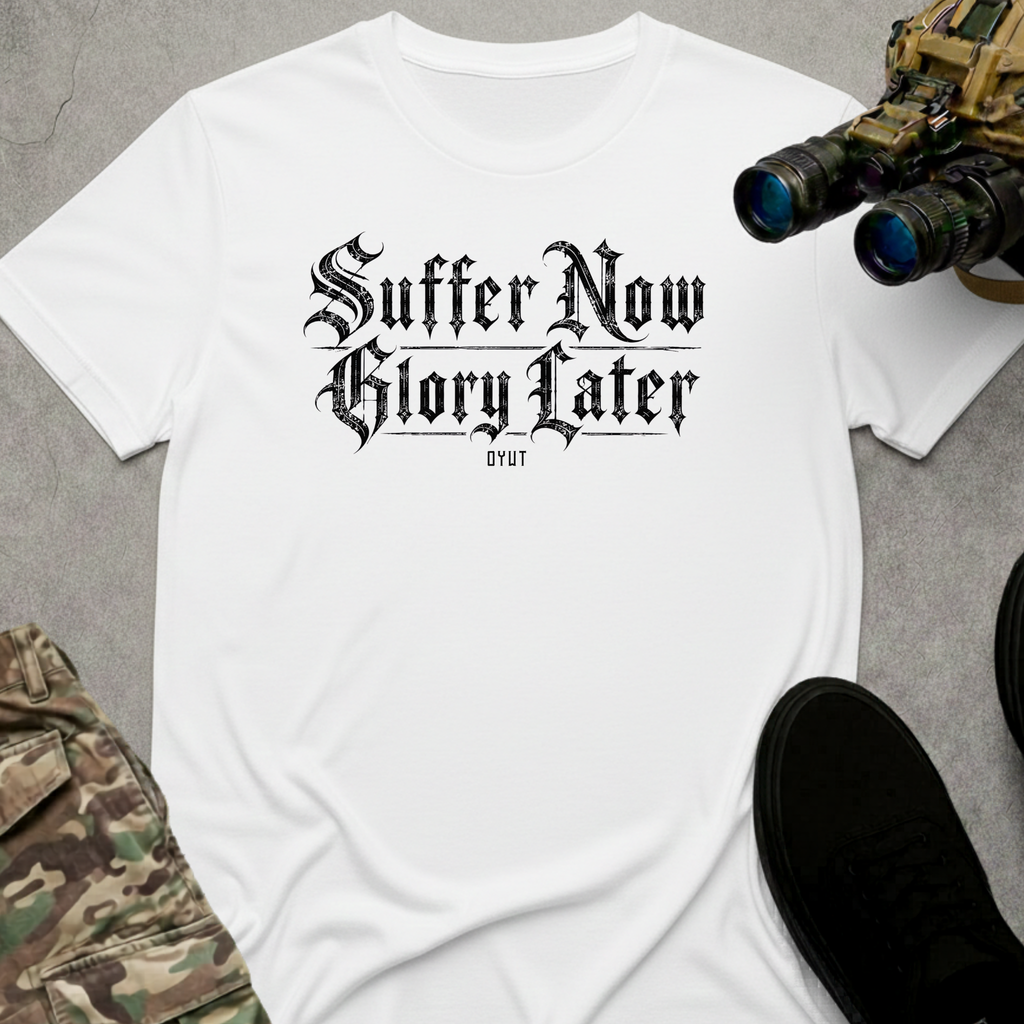 A white crewneck t-shirt featuring a black graphic print that reads 'Suffer Now Glory Later'. The shirt is styled in a flat-lay arrangement with tactical gear props, including binoculars, a black shoe, and camouflage pants, on a dark gray background.