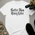 A white crewneck t-shirt featuring a black graphic print that reads 'Suffer Now Glory Later'. The shirt is styled in a flat-lay arrangement with tactical gear props, including binoculars, a black shoe, and camouflage pants, on a dark gray background.