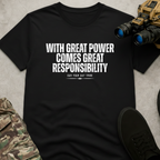 A black short-sleeve graphic T-shirt laid flat on a light gray surface, with bold white text reading “WITH GREAT POWER COMES GREAT RESPONSIBILITY” and smaller text “OUR YOUR DAY THRIE” beneath it. The shirt is styled with camouflage pants, black shoes, and binoculars around it.