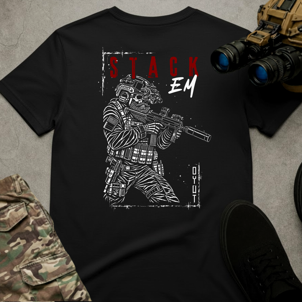 A black graphic t-shirt laid flat on a gray textured surface, featuring a white tactical soldier illustration and the text 'STACK EM' in red and white. The shirt is styled with camouflage pants, black sneakers, and tactical binoculars.