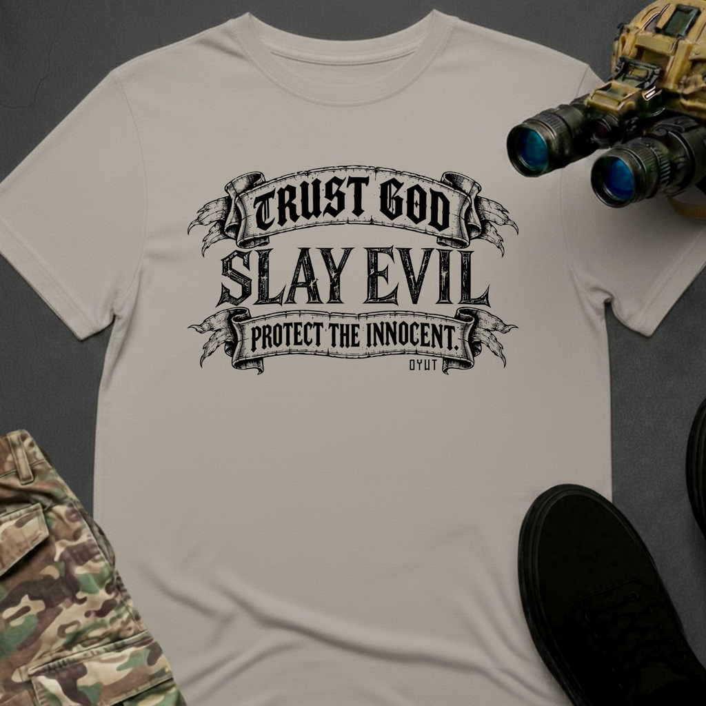 A light beige crewneck t-shirt featuring a black graphic print that reads 'TRUST GOD SLAY EVIL PROTECT THE INNOCENT' with 'GYOT' below. The shirt is styled in a flat-lay arrangement with tactical gear props, including binoculars, a black shoe, and camouflage pants, on a dark gray background.