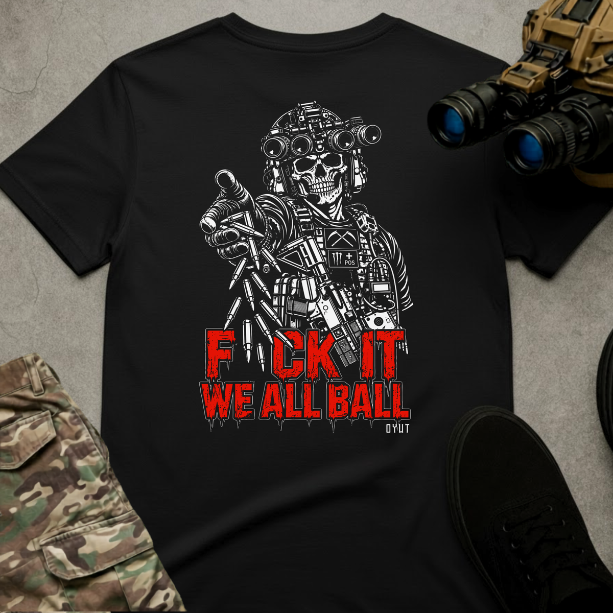 A black short-sleeve graphic T-shirt laid flat on a gray textured surface, surrounded by tactical-themed props including binoculars, camouflage pants, and a black shoe. The shirt features a large white illustrated soldier/skull graphic with red text reading 'F*CK IT WE ALL BALL' and a small 'DYOT' mark below.
