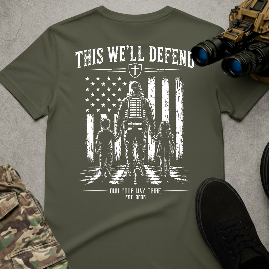 An olive green graphic t-shirt laid flat on a gray textured surface, surrounded by tactical props including binoculars, a toy vehicle, camouflage pants, and a black shoe. The shirt features a distressed white graphic of a soldier protecting two children in front of a stylized American flag, with the text 'THIS WE'LL DEFEND' and 'OUR WAY TRIBE EST.