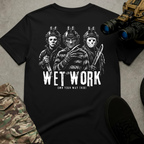 A black short-sleeve graphic T-shirt laid flat on a gray textured surface, surrounded by styled props including binoculars, a black shoe, camouflage fabric, and a helmet. The shirt features a large white horror-style graphic of three masked figures above the text 'WET WORK' and smaller text 'OWN YOUR WAY TRIBE'.